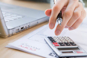 Person using calculator and pen on financial documents near a laptop.