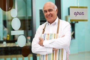 Smiling older man in a colorful apron with arms crossed.